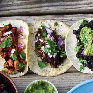 The image shows a group of tacos on a table from Dos Amigos food truck. The tacos seem to be a variety of Mexican and Tex-Mex food. Ingredients like guacamole, leaf vegetables, and corn tortillas are also included.