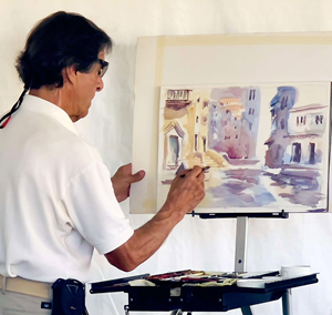 The image captures a man intently focused on painting a watercolor artwork. He is positioned in the foreground, facing left and slightly away from the viewer, engrossed in his creative process. The man has dark hair, pulled back loosely, and is wearing dark sunglasses. He is dressed in a white polo shirt with the collar popped. His left arm is holding the edge of the painting's paper, while his right hand holds a paintbrush, actively applying color to the canvas. His posture suggests concentration and dedic