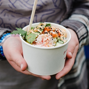 The image shows a person holding a paper bowl filled with a fresh, colorful poke bowl. The dish appears to include ingredients like shredded crab, diced salmon, cucumber, edamame, seaweed, and greens, topped with sesame seeds and a drizzle of sauce. A wooden utensil is sticking out from the bowl. The person is dressed in a patterned top and layered clothing, and they’re wearing beaded bracelets. The overall vibe suggests a casual, outdoor food market or festival setting.