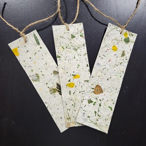 The image displays three handmade bookmarks, each made from a pale, off-white paper embedded with dried botanical elements. The bookmarks are arranged diagonally from left to right, slightly overlapping. They are rectangular in shape with rounded top corners and a single hole punched near the top center of each, through which a piece of rough, natural-colored twine is threaded and tied in a simple knot at the top. The twine extends upwards and out of frame.