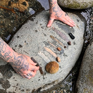 The image is a close-up, overhead shot focusing on a person's tattooed hands arranging small objects on a large, flat, grey, oval-shaped rock. The rock is wet, and the water appears to be shallow, surrounding the rock in places. The surface of the main rock is textured with small, dark speckles and has a somewhat rough appearance.