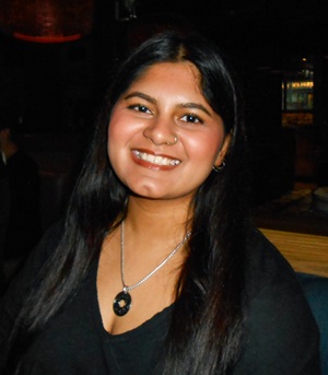The image is a medium shot portrait of a young Indian woman with dark, straight hair that falls past her shoulders. She is smiling broadly, her head tilted slightly to her left. Her eyes are dark and appear to be looking slightly off-camera. She is wearing a dark, possibly black, V-neck top and a silver necklace with a round, dark pendant featuring an intricate design. She also has a nose ring and small hoop earrings. Her skin has a warm, tan tone.