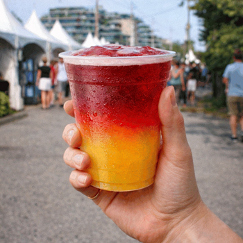 The image is a close-up, eye-level shot of a person's hand holding a clear plastic cup filled with a layered frozen drink. The drink is divided into two distinct layers: the bottom layer is a bright, vibrant yellow, and the top layer is a rich, deep red. 