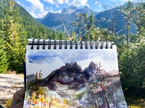 The image shows a hand holding an open sketchbook, displaying a watercolor painting of a mountain landscape. The sketchbook is bound with black spiral wire. The painting itself depicts a dramatic mountain range with sharp, jagged peaks rendered in dark grays, purples, and blacks, suggesting shadows and rock formations. Some lighter tones indicate highlights and snow patches on the upper slopes.