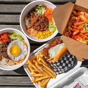 A variety of Korean and Canadian takeout foods are laid out on a wooden surface. The spread includes two bowls of bibimbap, a portion of poutine, and a burger with fries.