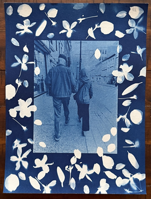 The image is a cyanotype print featuring a central photograph surrounded by a decorative border of botanical elements. The overall color palette is dominated by deep blues and crisp whites, characteristic of the cyanotype process.