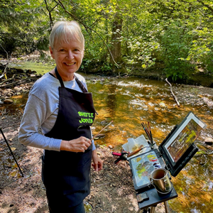 The image captures a joyful outdoor painting scene set beside a shallow, sun-dappled stream. In the foreground, a woman with short, light blonde hair and a warm smile stands to the left of the frame. She is wearing a long-sleeved grey top and a black apron with the words "SKYLE JONES" emblazoned in bright green lettering. She holds a paintbrush in her right hand and appears to be mid-stroke or about to apply paint.