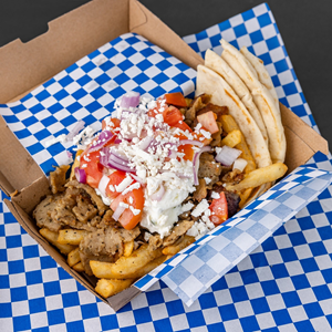 The image showcases a delicious-looking Greek food dish, likely a gyro or similar platter, presented in a cardboard takeout container. The container is lined with a vibrant blue and white checkered paper, creating a bold and eye-catching background
