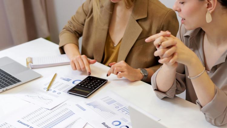 Two people sit at a desk with a laptop, paper files, and a calculator.