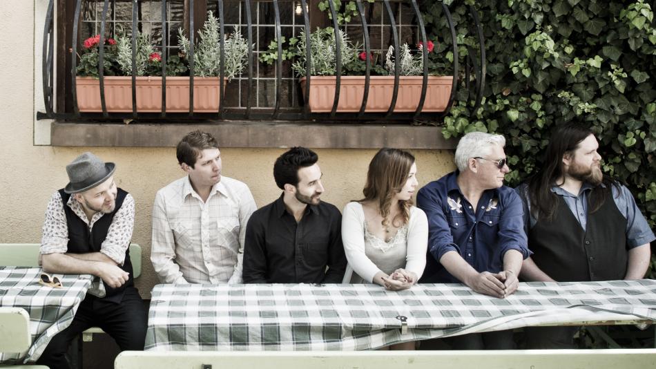 The image displays a group of six individuals, three men and three women, seated in a row at an outdoor table, likely a cafe or restaurant patio. They are all looking to their right, creating a unified gaze that draws the viewer's attention in that direction. The scene is set against a neutral beige wall and a window with decorative bars above it. Two terra-cotta planters filled with greenery and a few red flowers are positioned on a ledge beneath the window. To the right of the window, a dense wall of lush