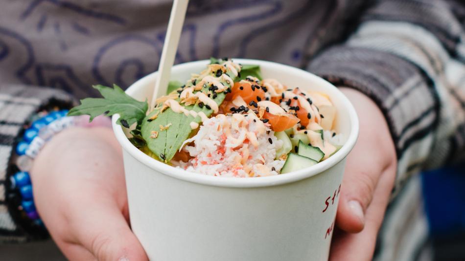 The image shows a person holding a paper bowl filled with a fresh, colorful poke bowl. The dish appears to include ingredients like shredded crab, diced salmon, cucumber, edamame, seaweed, and greens, topped with sesame seeds and a drizzle of sauce. A wooden utensil is sticking out from the bowl. The person is dressed in a patterned top and layered clothing, and they’re wearing beaded bracelets. The overall vibe suggests a casual, outdoor food market or festival setting.