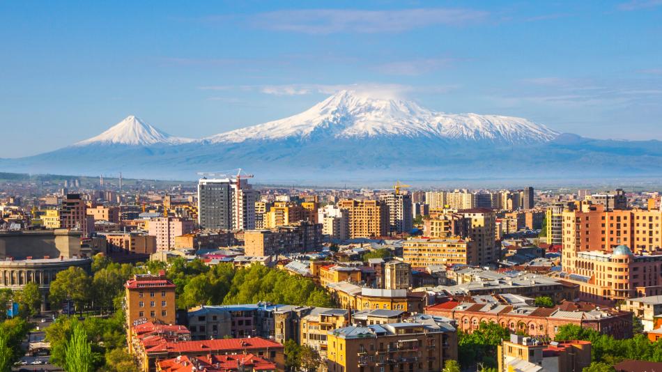 The image depicts a city with a backdrop of Mount Ararat, towering at 5,137 meters. The mountain is snow-capped and features two major volcanic cones. This view is from Yerevan, Armenia, set against a landscape that includes buildings, trees, and a cloudy sky. The scene combines urban elements with the natural beauty of the mountain and surrounding nature.