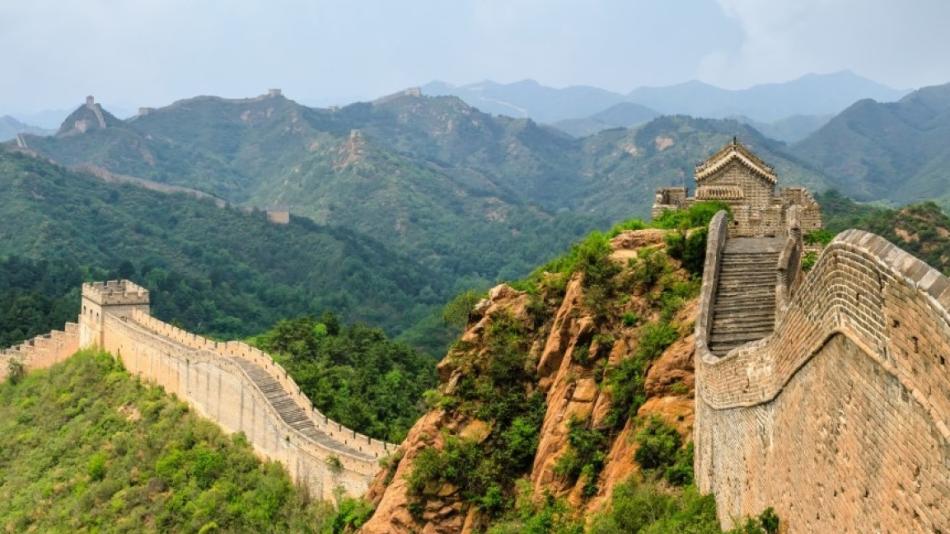 The image is of a stone wall on a hill, part of the Great Wall of China. The setting is outdoors with clouds and sky in the background, surrounded by a landscape with trees and mountains.