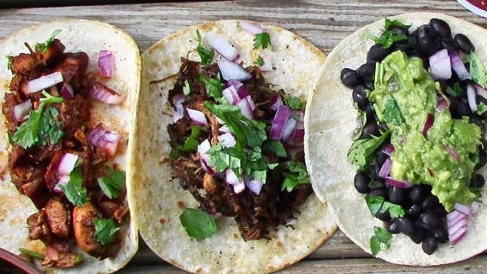 The image shows a group of tacos on a table from Dos Amigos food truck. The tacos seem to be a variety of Mexican and Tex-Mex food. Ingredients like guacamole, leaf vegetables, and corn tortillas are also included.