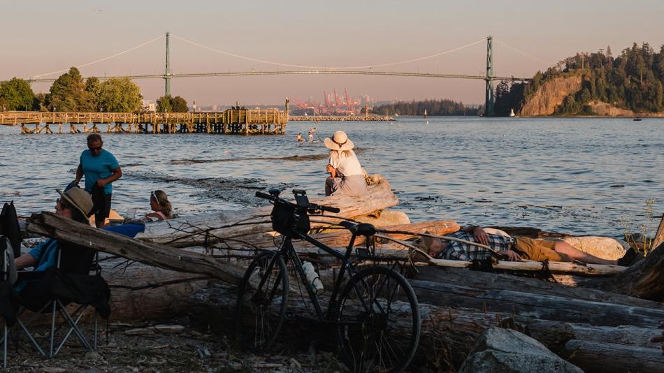 A woman sits on a log by the water, with a bicycle nearby, surrounded by a group of people enjoying the scenery.