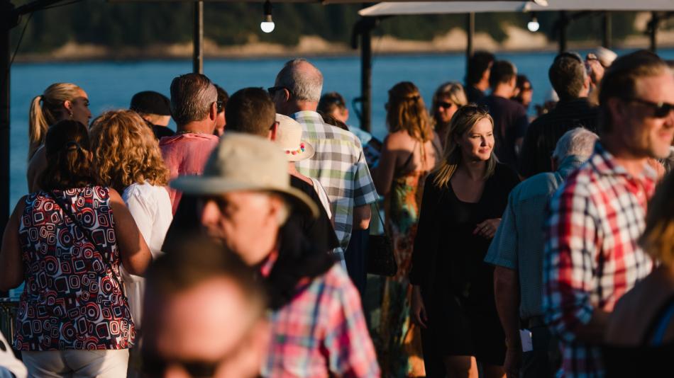 Full shot of a large crowd of people, likely at an outdoor event or gathering by a body of water.   The people are densely packed together, with various ages and styles of clothing.   Many people are wearing casual attire, including shirts, tops, and dresses, and some are visible with hats and sunglasses. 