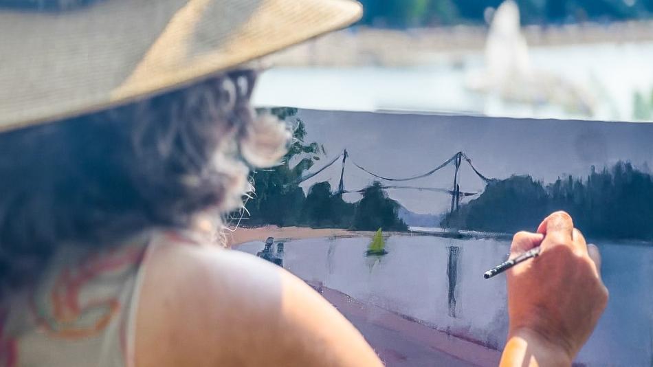 A woman is outdoors, painting a picture while wearing a stylish sun hat. The scene captures her artistic expression during the summer. She embodies a sense of fashion and creativity, blending art with her personal style.