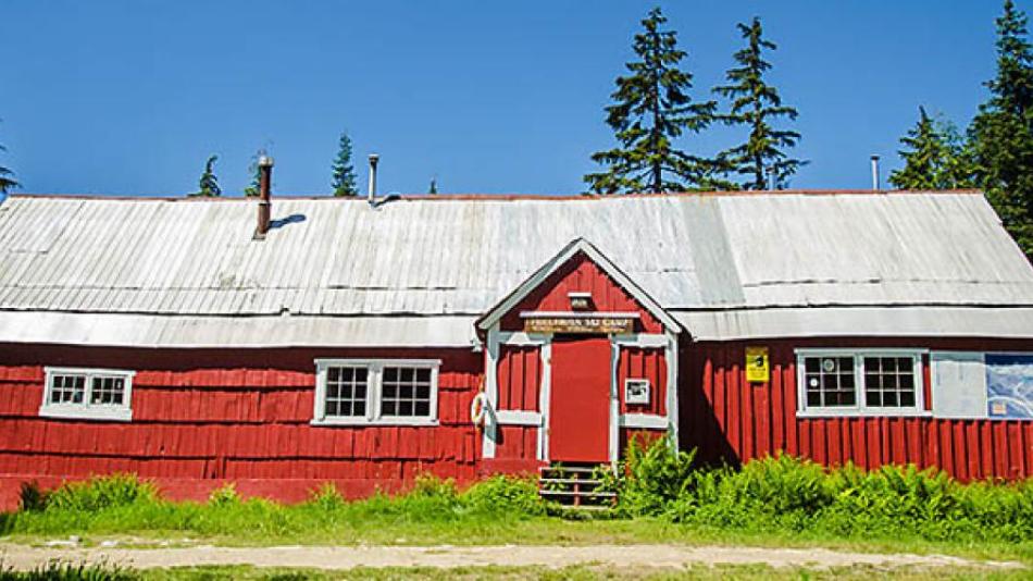 A long, red barn-like building in front of a forest of evergreen trees.