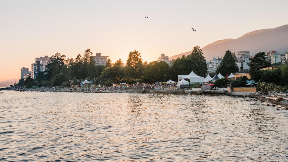 The image captures a scenic view of an event taking place along a shoreline, possibly a festival or outdoor gathering, with a body of water in the foreground. In the background, there are buildings, trees, and what appears to be a mountain range partially obscured by the hazy sunlight. The sky is a soft gradient of colors, suggesting either sunrise or sunset, and two birds are seen flying overhead. 