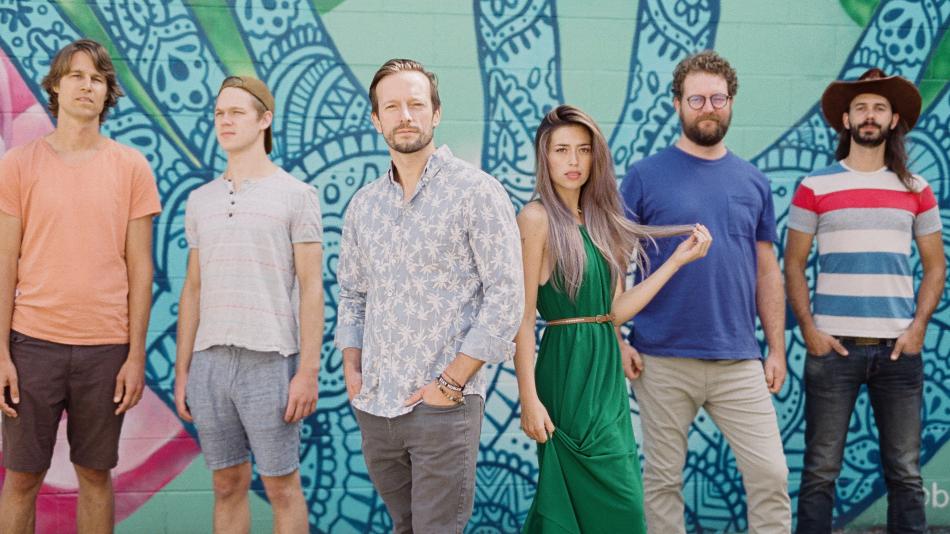 The image shows a group of six people standing side-by-side in front of a vibrant, colorful mural. The mural features intricate blue and green patterns that resemble mandala or floral designs, adding a striking artistic backdrop.