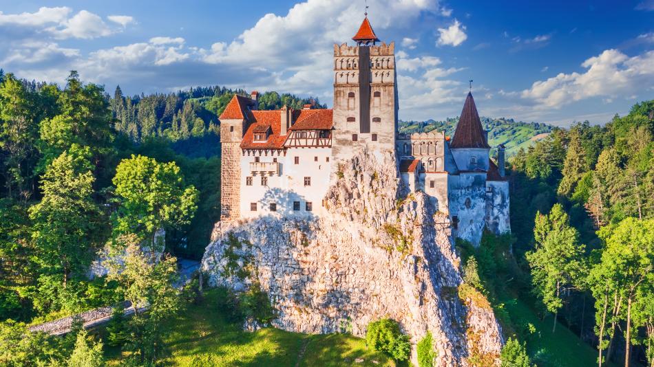 This image shows Bran Castle, a dramatic medieval fortress perched on a rocky hilltop, surrounded by dense green forest under a bright blue sky with scattered clouds. The castle features tall towers with red-tiled roofs and stone walls, giving it a classic Gothic appearance. Bran Castle is famously associated with the Dracula legend and is located in Transylvania, Romania. The lush greenery and mountainous landscape create a picturesque and mysterious setting.