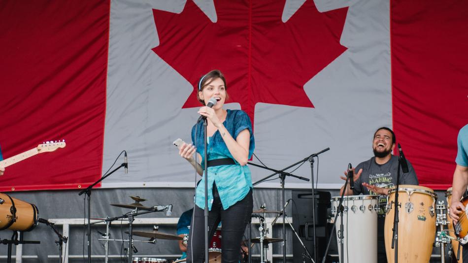 A diverse group of musicians performs on stage, featuring a female singer, male guitarist, bass player, and drummer, with a Canadian flag.