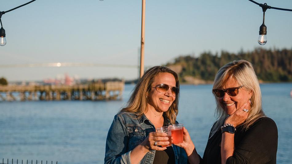 The image depicts two women in their 40s or 50s standing outdoors near a body of water, likely enjoying a summer evening. They are positioned in the foreground, with their upper bodies and faces prominently displayed. Both women are smiling and appear to be in conversation, holding clear plastic cups with reddish-pink drinks.