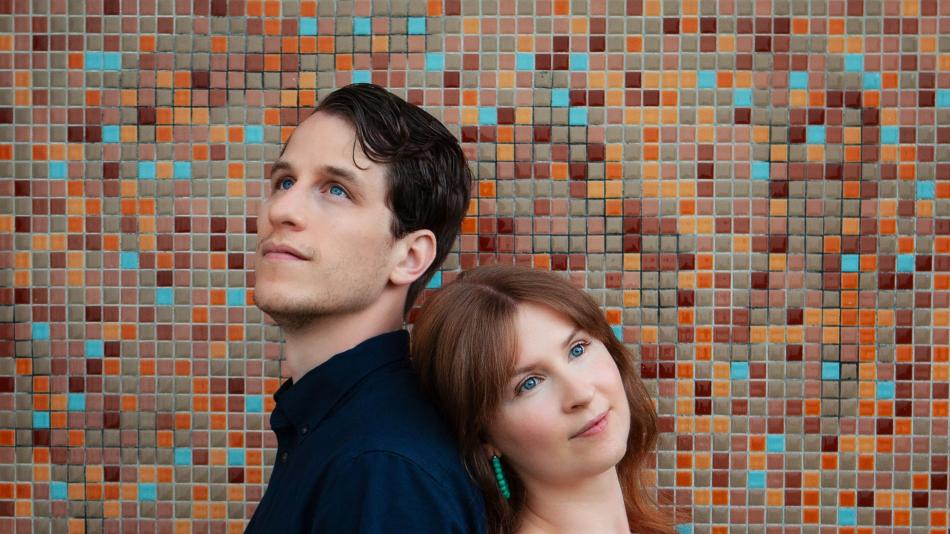 The image is a medium shot portrait of a man and a woman positioned back-to-back against a colorful, mosaic tiled wall. The man is on the left, facing upwards and slightly to the left, with his head tilted back. He has short, dark brown hair, fair skin, and striking blue eyes. He is wearing a dark navy blue collared shirt.