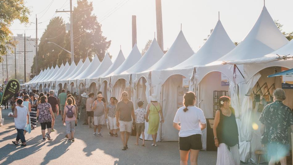 A bustling art market scene with a large group of people strolling along a street filled with colorful tents.