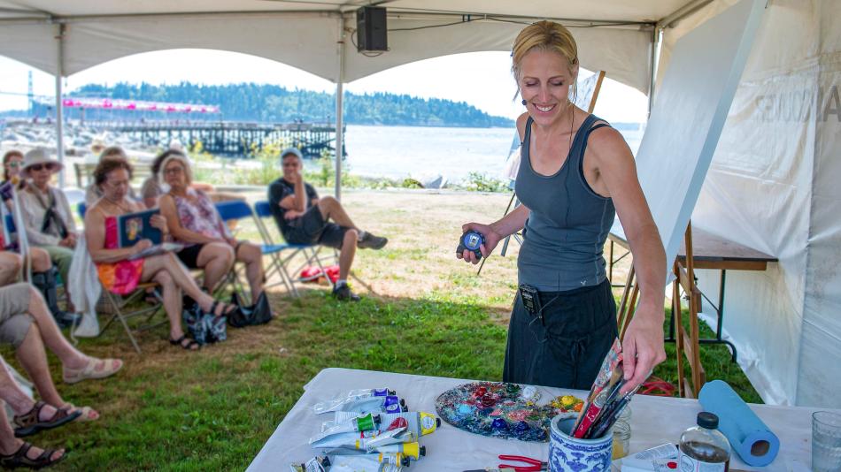 A woman is standing next to a table filled with art supplies. The setting appears to be outdoors, possibly during summer, surrounded by grass and a picnic atmosphere. There are also other people present in the scene, including a man and a group engaged in activities around a tent.