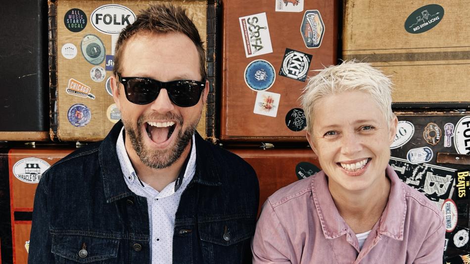 The image is a medium shot of a man and a woman, both smiling broadly and looking at the camera. They are positioned side-by-side in front of a wall of vintage suitcases.