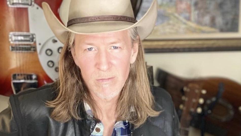 The image is a medium shot portrait of a middle-aged man with long, blonde, wavy hair, wearing a light brown cowboy hat. He has a serious expression and is looking directly at the camera. He is wearing a dark leather jacket over a blue and white plaid shirt.