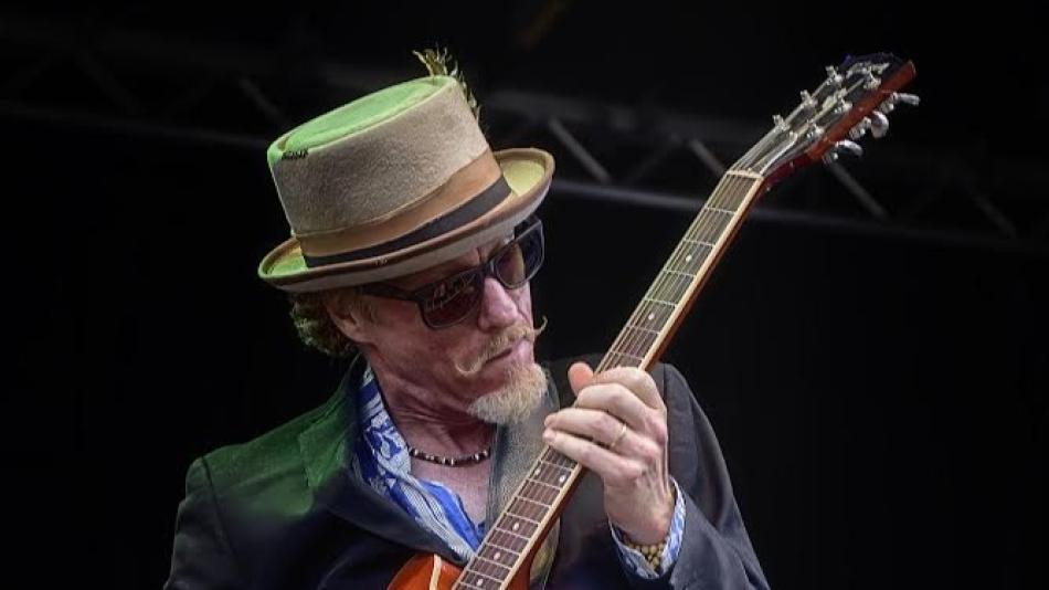 The image captures a medium close-up of a male musician playing a guitar on a stage, likely during a performance. The background is dark, suggesting an indoor venue or an outdoor event at night, with some stage lighting faintly visible in the upper part of the frame.
