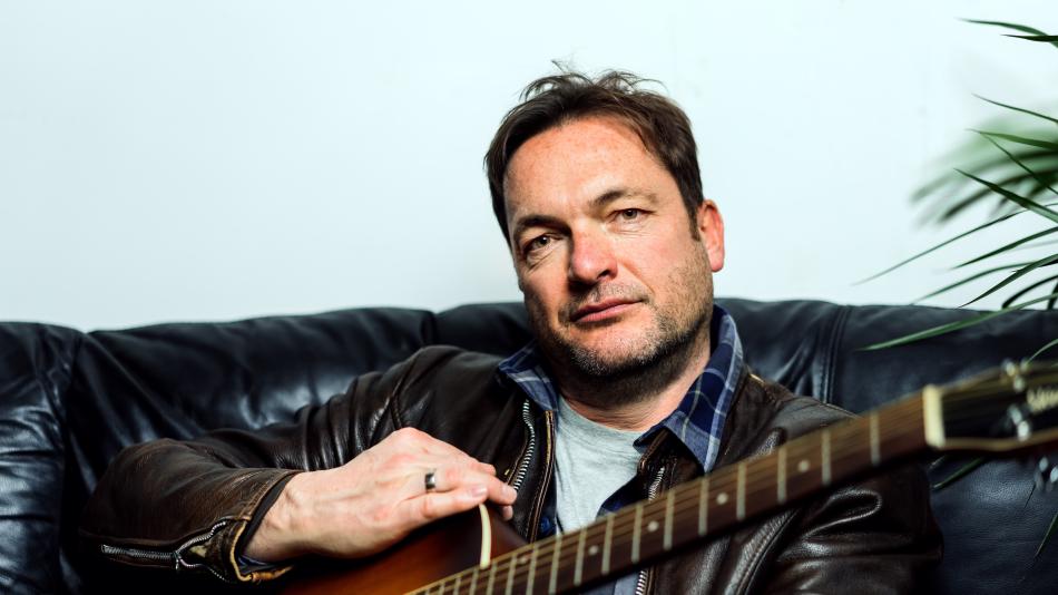 A medium shot portrait features a middle-aged man with fair skin, light brown hair, and a slight stubble. He is seated on a black leather couch, facing directly towards the camera with a neutral, contemplative expression. His eyes, a light brown color, are looking straight ahead. He has a few freckles visible on his cheeks and forehead.