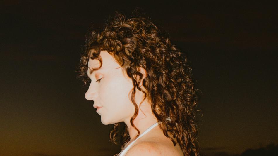 The image is a close-up, profile view of a young woman with voluminous, dark curly hair. Her head is tilted downwards, and her eyes are closed, suggesting a moment of reflection or peace. Her skin tone is fair, and she appears to be wearing subtle makeup, with a hint of eyeshadow. A delicate silver necklace is visible around her neck, and the top of a white garment, possibly a tank top or dress, can be seen.