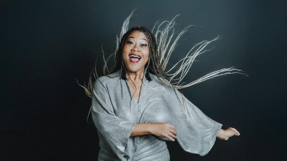 A woman is captured in a dynamic, mid-movement shot against a dark, neutral background. She appears to be of African descent and is wearing a shimmering, silver, V-neck dress with wide, flowing sleeves. Her expression is joyful and energetic, with her mouth open as if she is laughing or exclaiming. Her eyes are wide and her makeup is dramatic, with bold eyeliner and bright red lipstick.
