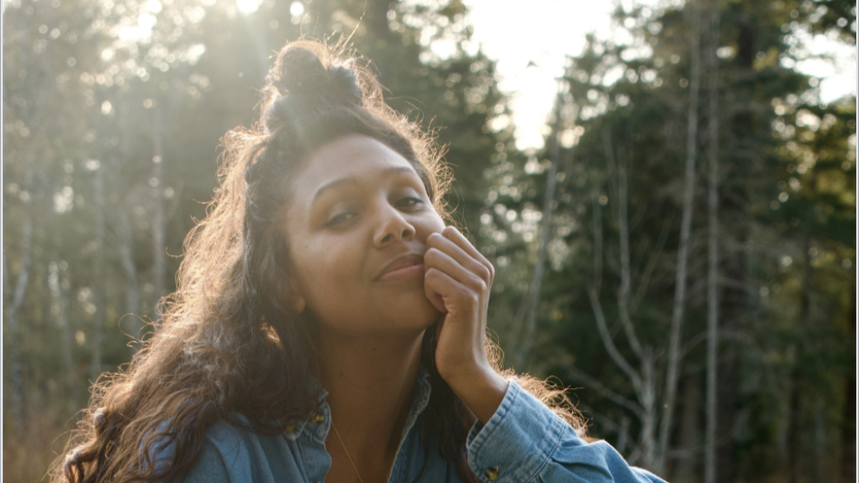 The image is a medium shot portrait of a young woman with dark, curly hair, some of which is pulled up into a messy bun at the top of her head. She is looking towards the camera with a gentle, slightly upward gaze and a soft smile playing on her lips. Her right hand is brought up to her face, with her fingers resting lightly against her cheek and chin, a pose that suggests contemplation or contentment.