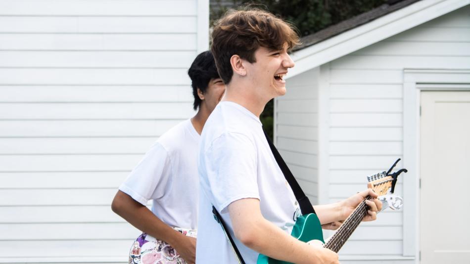 The image captures two young men outdoors, positioned in front of a white clapboard-sided building, likely a residential structure. The composition is dynamic, with the primary subject—a young man with tousled brown hair—occupying the right side of the frame. 