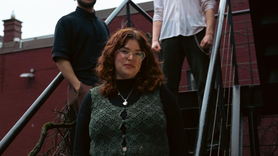 The image is an outdoor, medium shot of three people standing on a metal staircase against a brick building. The central figure is a young woman with curly, reddish-brown hair, wearing glasses with thin, gold-toned frames. She has fair skin and is looking directly at the camera with a slight smile. She is wearing a dark turtleneck shirt and a distinctive, dark green crocheted vest with a floral pattern and buttons down the front. A delicate silver necklace with a locket-shaped pendant hangs around her neck