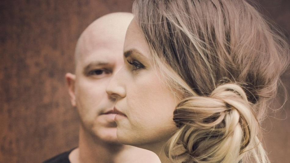 The image is a close-up, profile shot of two people, a man and a woman, superimposed or layered in a way that suggests a connection or duality. The man is bald and in the background, seen from the nose up, facing left. His expression is neutral. The woman is in the foreground, facing left, with her head angled slightly downwards. Her hair is light brown with blonde highlights, pulled back into a messy bun at the nape of her neck. Her makeup is visible, with eyeliner and mascara accentuating her eyes. 