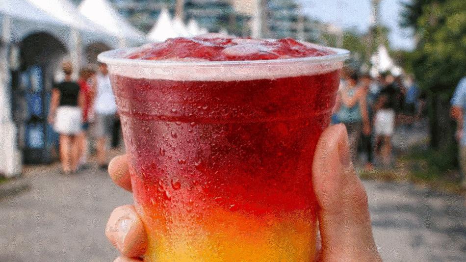 The image is a close-up, eye-level shot of a person's hand holding a clear plastic cup filled with a layered frozen drink. The drink is divided into two distinct layers: the bottom layer is a bright, vibrant yellow, and the top layer is a rich, deep red. 