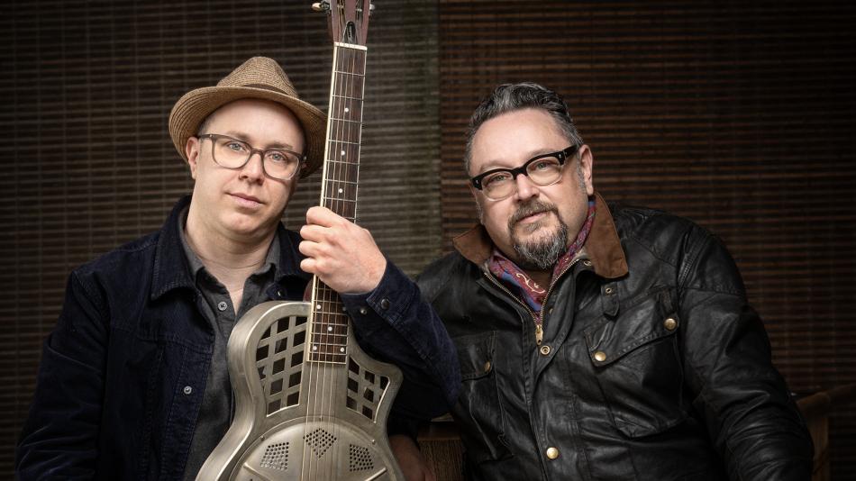 The image is a medium shot portrait of two middle-aged men, positioned side-by-side and looking towards the camera. They are both wearing glasses and appear to be musicians, as one of them is holding a resonator guitar.