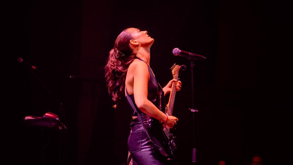 The image captures a dynamic moment of a female musician performing on stage. She is positioned in the center-left of the frame, looking upwards with her head tilted back, her eyes closed, exuding a sense of passionate immersion in her performance. Her dark, wavy hair is tied back into a ponytail and flows loosely around her shoulders and back.