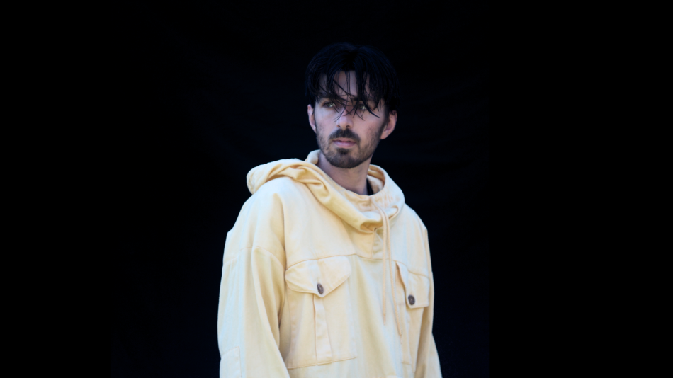 A man with dark, somewhat messy hair and a beard is shown from the chest up, looking to his left with a neutral expression. He is wearing a light yellow, oversized hooded jacket with a drawstring and two large buttoned pockets on the front. The jacket appears to be made of a sturdy material like canvas or denim. The background is completely black and featureless, creating a stark contrast with the man and his clothing. The lighting highlights the textures of the fabric and the contours of the man's face and
