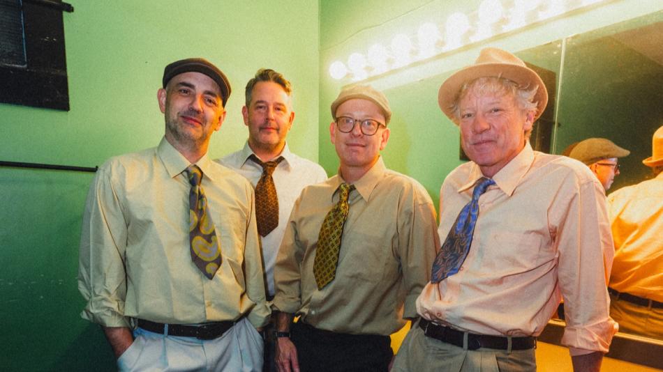The image features a group of four middle-aged men, dressed in retro-style attire, posing for a photograph. They are all wearing light-colored button-down shirts and ties, with variations in their hats and pants. The man on the far left wears a flat cap and a tan shirt with a colorful tie featuring a paisley-like pattern in yellow, grey, and brown. He also sports light grey pleated pants and a dark belt, with his left hand in his pocket. The second man from the left wears a dark patterned tie and a white sh