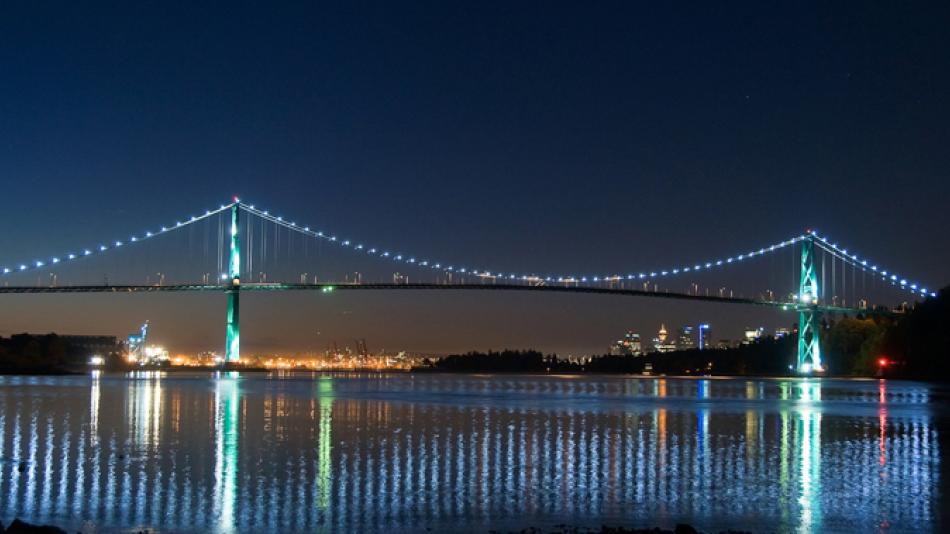 The Lions Gate Bridge seen at night with lights along the suspension cables. The water below reflect the bridges lights. 
