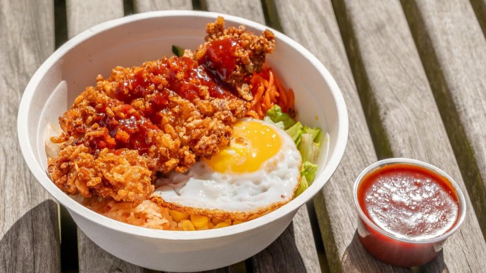 A white bowl filled with rice, fried chicken, a sunny-side-up egg, and shredded carrots sits on a wooden surface. A small cup of red sauce is placed beside the bowl.