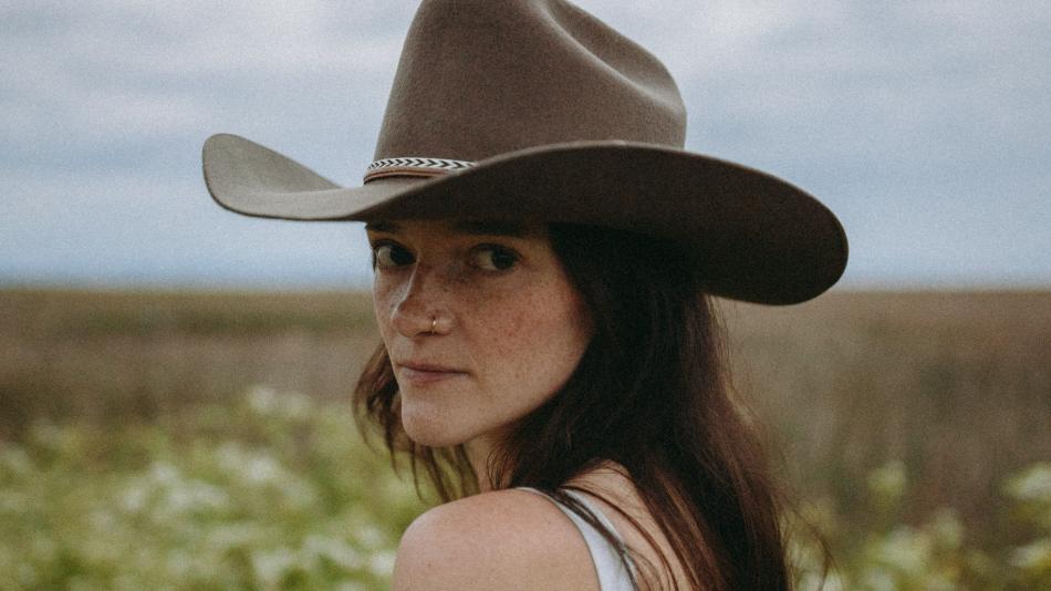 The image is a close-up portrait of a young woman wearing a wide-brimmed, dark brown cowboy hat. She is looking over her left shoulder towards the camera, with her face angled slightly to the right. Her expression is calm and direct, with a hint of a gaze.