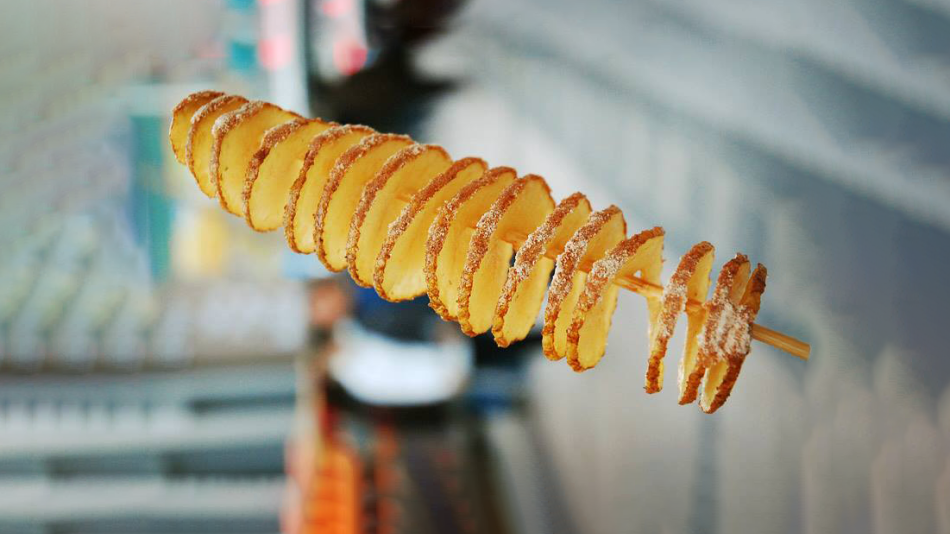 The image features a close-up, vertical shot of a spiral-cut potato, skewered and standing upright. The potato slices are thin and circular, arranged in a helix around a wooden skewer. Each slice is golden brown and appears to be lightly seasoned, with a dusting of white seasoning, possibly salt or sugar, visible along the edges. The texture of the potato looks crispy and slightly oily.