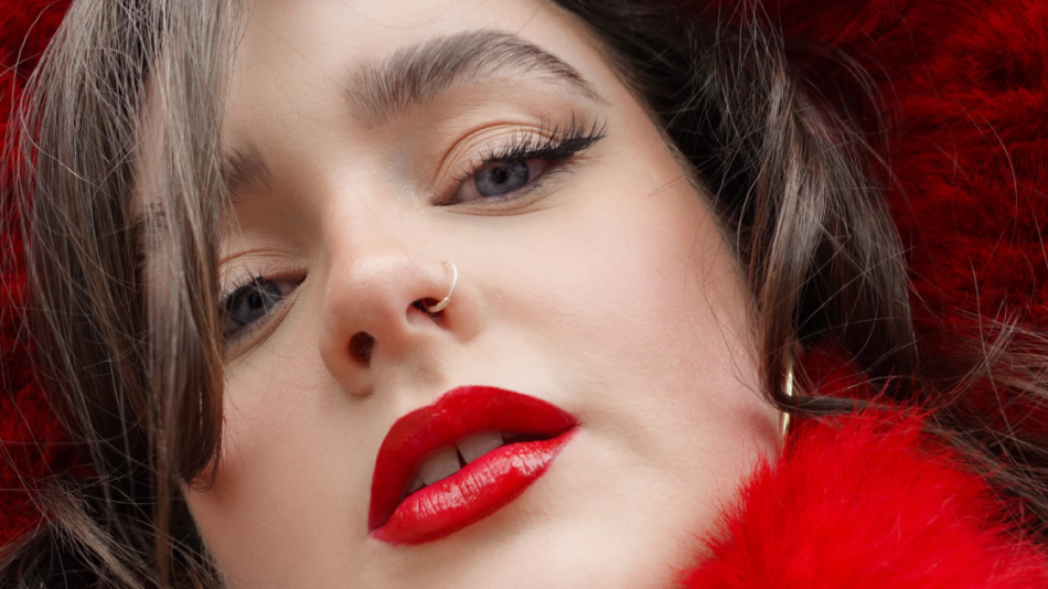 The image is a close-up, low-angle portrait of a young woman's face, with her head tilted slightly upwards and to the left. The foreground is dominated by her face, with the background largely obscured by vibrant, fluffy red material, possibly faux fur trim or a scarf.